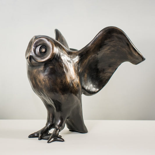 A bronze sculpture of an owl standing and looking forward with its wings outstretched behind it. The colour is a warm metallic black. The style is very curvy with smooth lines