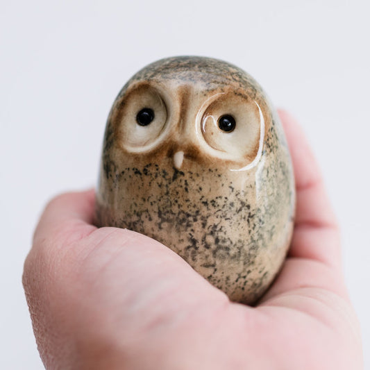 A small ceramic owl figurine with a speckled beige and brown glaze, held in a person's hand against a plain light background.