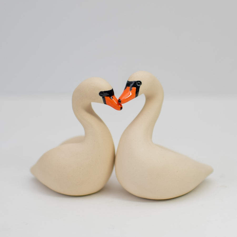 A pair of small white ceramic swan sculptures touching beaks on a plain background