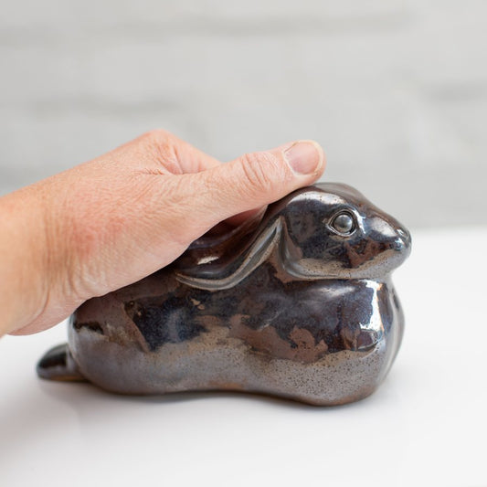 A ceramic hare sculpture with a glossy, metallic brown and silver glaze, resting on a white surface against a light gray brick wall background with a hand