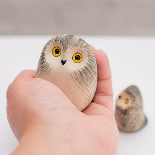 A hand holds a small, rounded owl figurine with yellow eyes and a black beak, with a second owl figurine blurred in the background on a white surface.
