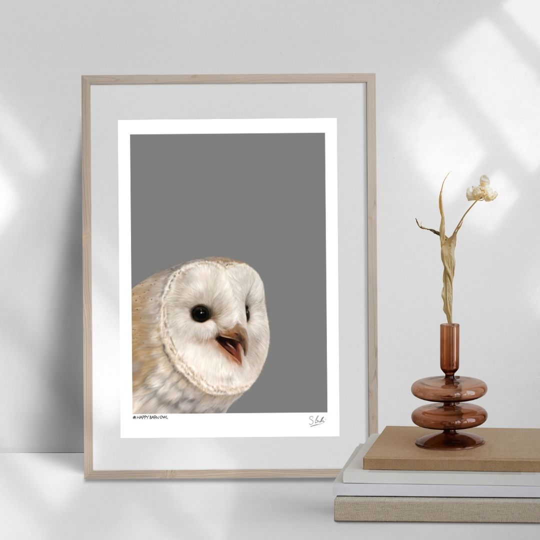 A framed art print featuring a close-up portrait of a barn owl on a gray background, styled leaning against a wall next to a decorative wooden vase with dried stems.
