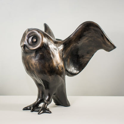 A bronze sculpture of an owl standing and looking forward with its wings outstretched behind it. The colour is a warm metallic black. The style is very curvy with smooth lines