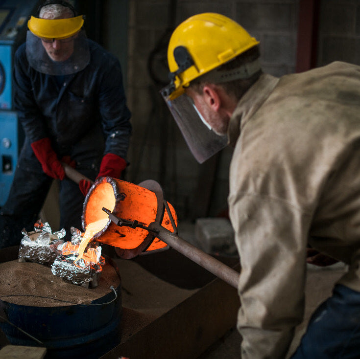 Two workers in protective gear pouring molten metal into molds