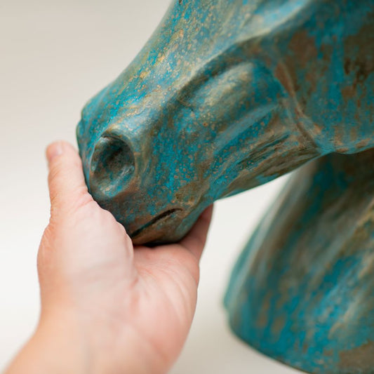 Hand touching the nose of a bronze horse head sculpture with a blue-green patina