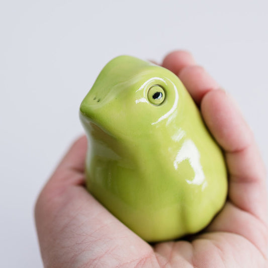 A glossy ceramic frog sculpture in a light lime green color, photographed against a neutral light gray background in a hand
