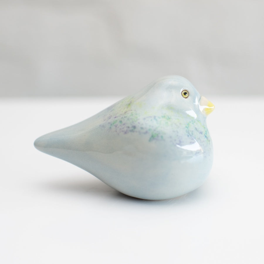 A small ceramic Pigeon figurine with a glossy pale blue finish, subtle green and purple speckled accents, and a yellow beak, photographed on a clean white background.