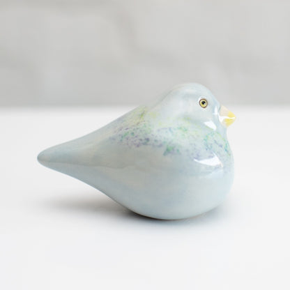 A small ceramic Pigeon figurine with a glossy pale blue finish, subtle green and purple speckled accents, and a yellow beak, photographed on a clean white background.