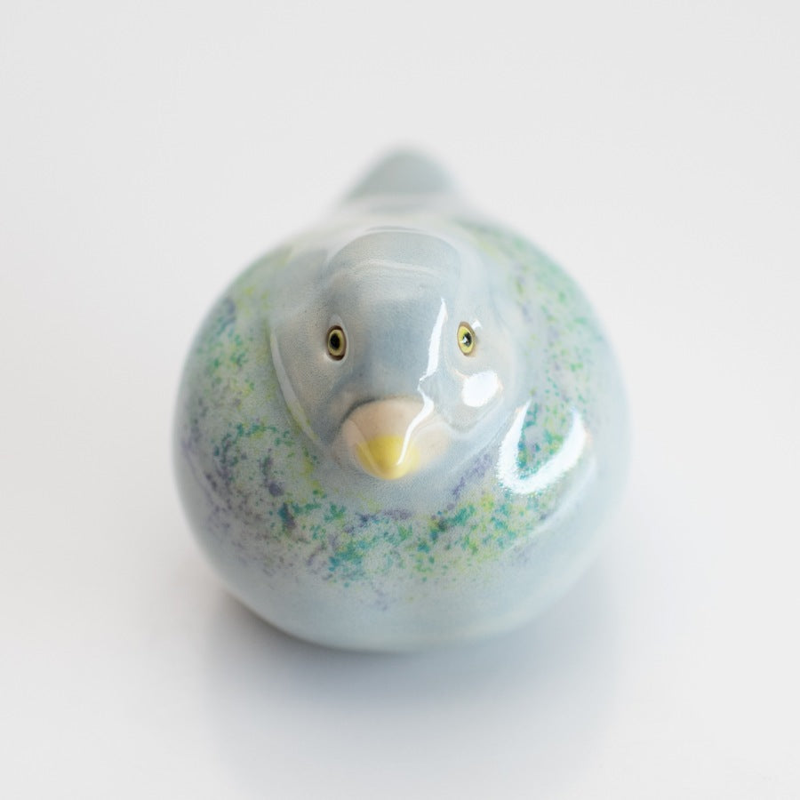 A small ceramic Pigeon figurine with a glossy pale blue finish, subtle green and purple speckled accents, and a yellow beak, photographed on a clean white background.