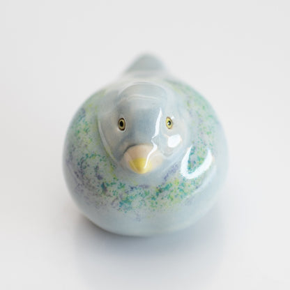 A small ceramic Pigeon figurine with a glossy pale blue finish, subtle green and purple speckled accents, and a yellow beak, photographed on a clean white background.