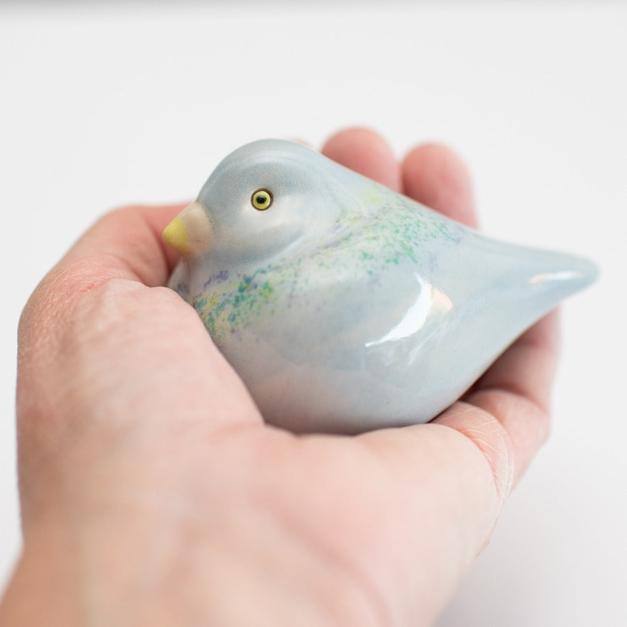 A small ceramic Pigeon figurine with a glossy pale blue finish, subtle green and purple speckled accents, and a yellow beak, photographed on a clean white background.
