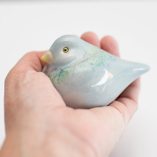 A small ceramic Pigeon figurine with a glossy pale blue finish, subtle green and purple speckled accents, and a yellow beak, photographed on a clean white background.