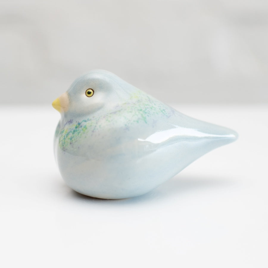 A small ceramic Pigeon figurine with a glossy pale blue finish, subtle green and purple speckled accents, and a yellow beak, photographed on a clean white background.