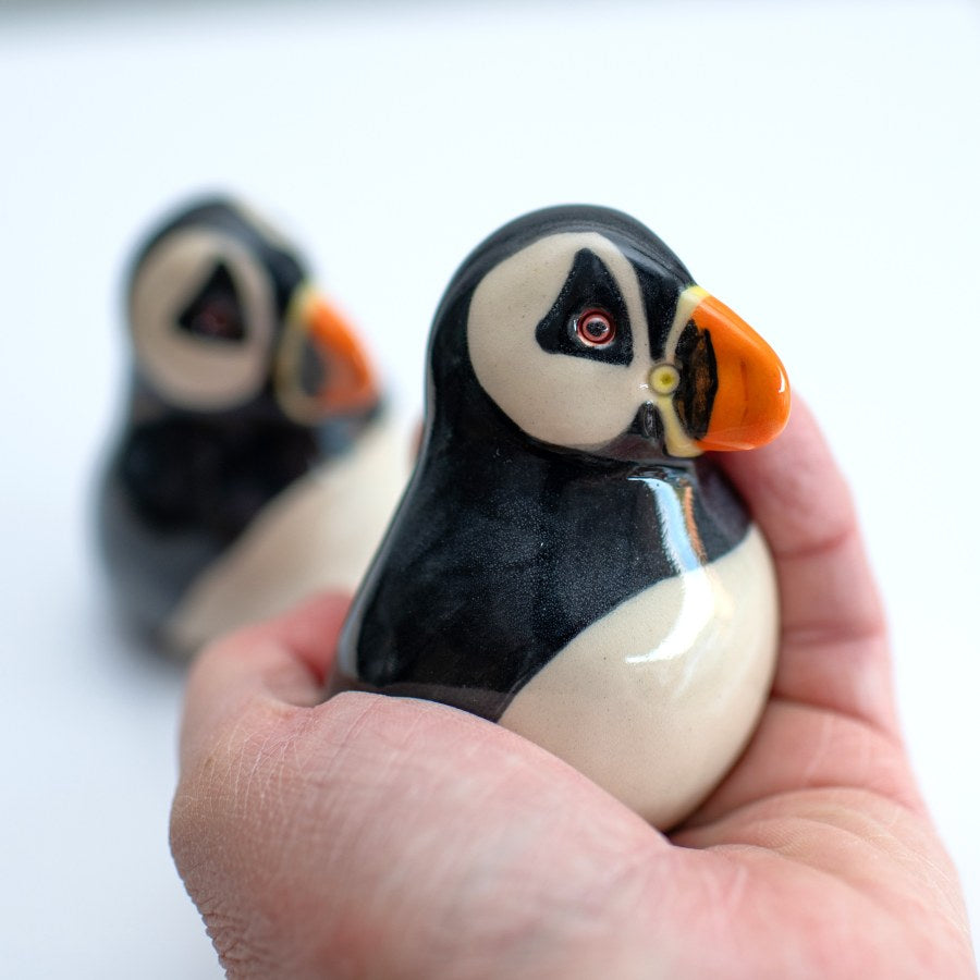 A hand holding a glossy ceramic puffin figurine, with a second blurred puffin in the background against a clean white backdrop.