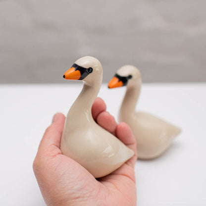 Hand holding a ceramic swan figurine with another swan in the background on a light surface.