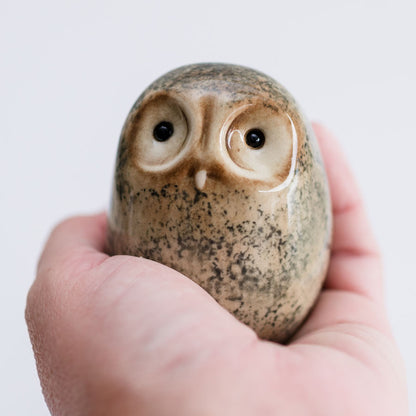 A small ceramic owl figurine with a speckled beige and brown glaze, held in a person's hand against a plain light background.