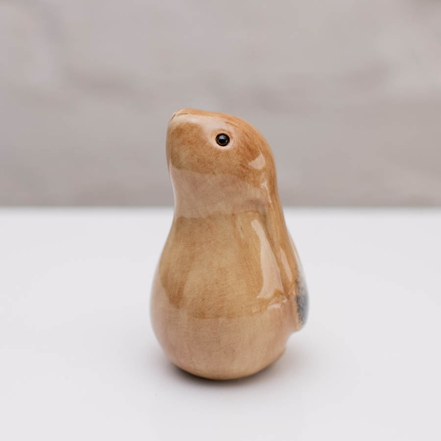 A small glazed ceramic hare figurine in warm tan and light brown tones, standing on a white surface against a soft gray background.