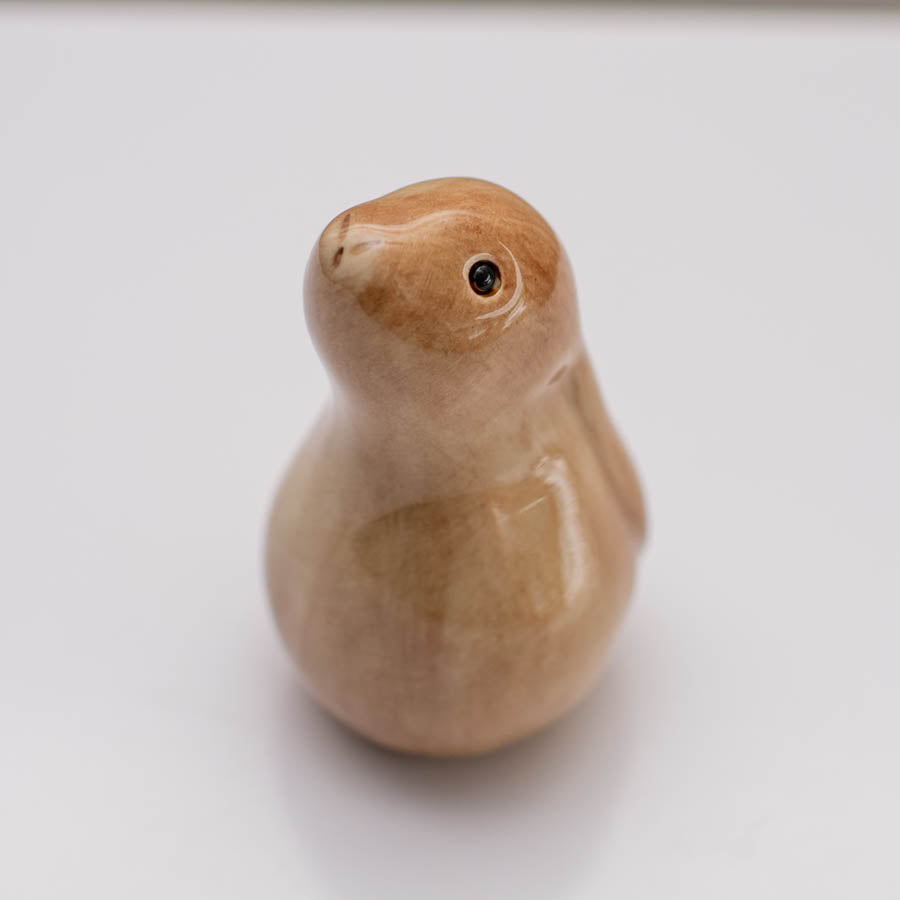 A small glazed ceramic hare figurine in warm tan and light brown tones, standing on a white surface against a soft gray background.