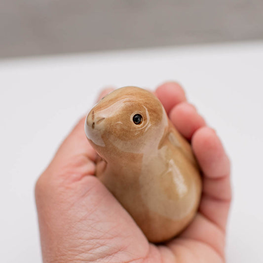 A small glazed ceramic hare figurine in warm tan and light brown tones, standing on a white surface against a soft gray background in a hand