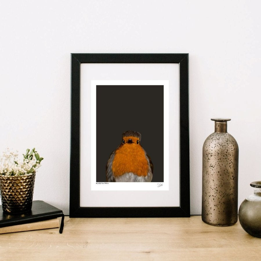 A framed art print of a robin bird against a dark background, styled on a wooden surface with decorative vases and a small plant.