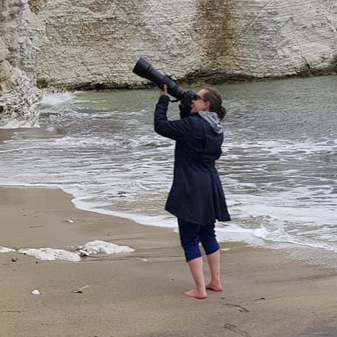Woman in dark coat and sunglasses holding a large camera lens on a sandy beach with waves