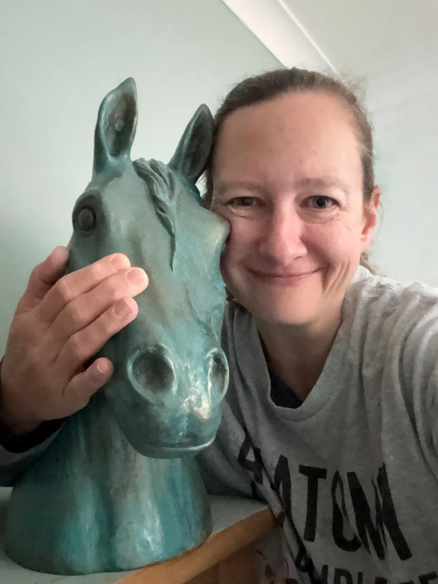Sarah smiling while holding a painted horse head sculpture with a distressed teal finish. She is wearing a grey graphic t-shirt and posing indoors against a light-colored wall.