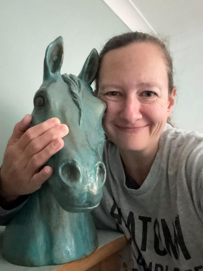 Sarah smiling while holding a painted horse head sculpture with a distressed teal finish. She is wearing a grey graphic t-shirt and posing indoors against a light-colored wall.