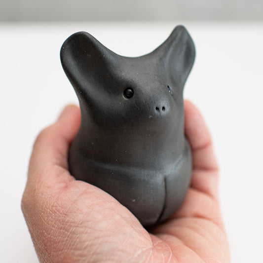 A matte black ceramic bat figurine standing upright on a white surface against a light gray background in a hand