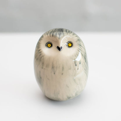 A small owl figurine with a glossy finish, featuring a cream-colored body with gray and black feather-like markings and bright yellow eyes, set against a neutral light gray background.