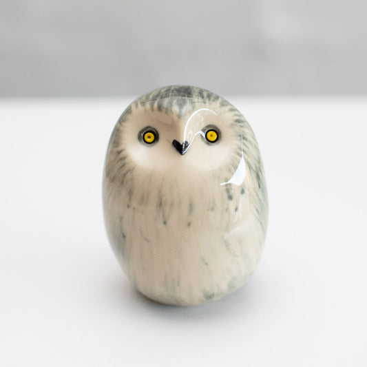 A small owl figurine with a glossy finish, featuring a cream-colored body with gray and black feather-like markings and bright yellow eyes, set against a neutral light gray background.
