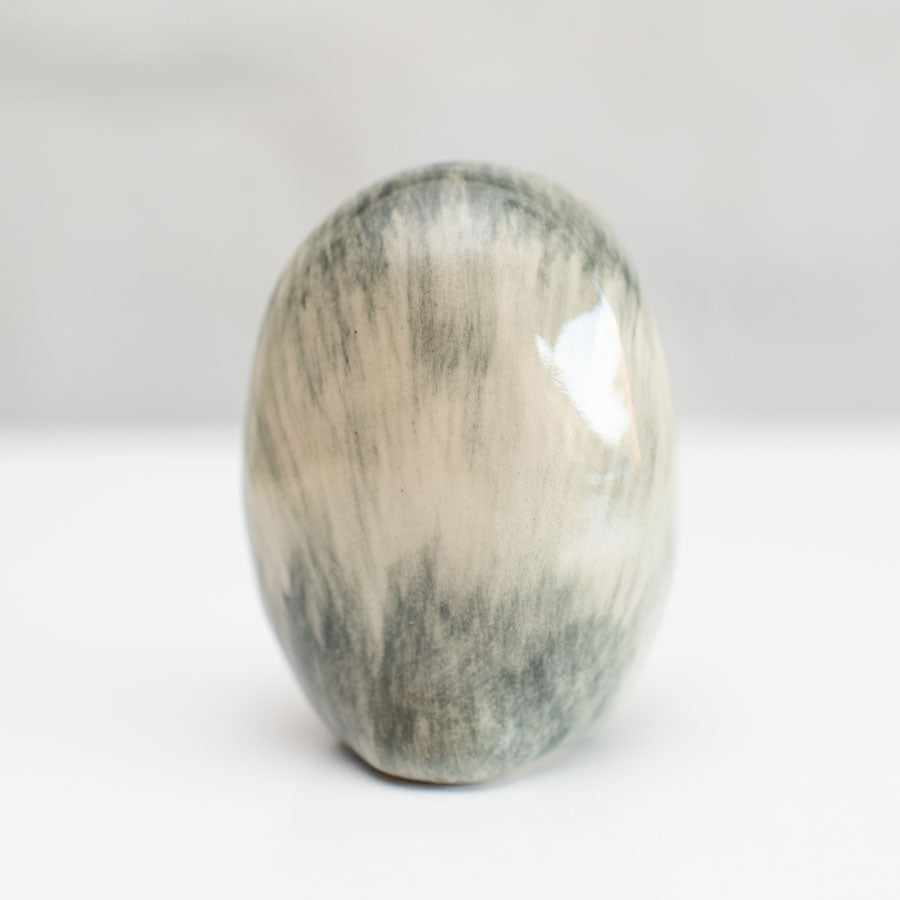 A polished oval stone with a marbled pattern of gray, beige, and white, standing upright on a white surface against a soft gray background.