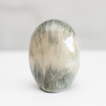 A polished oval stone with a marbled pattern of gray, beige, and white, standing upright on a white surface against a soft gray background.