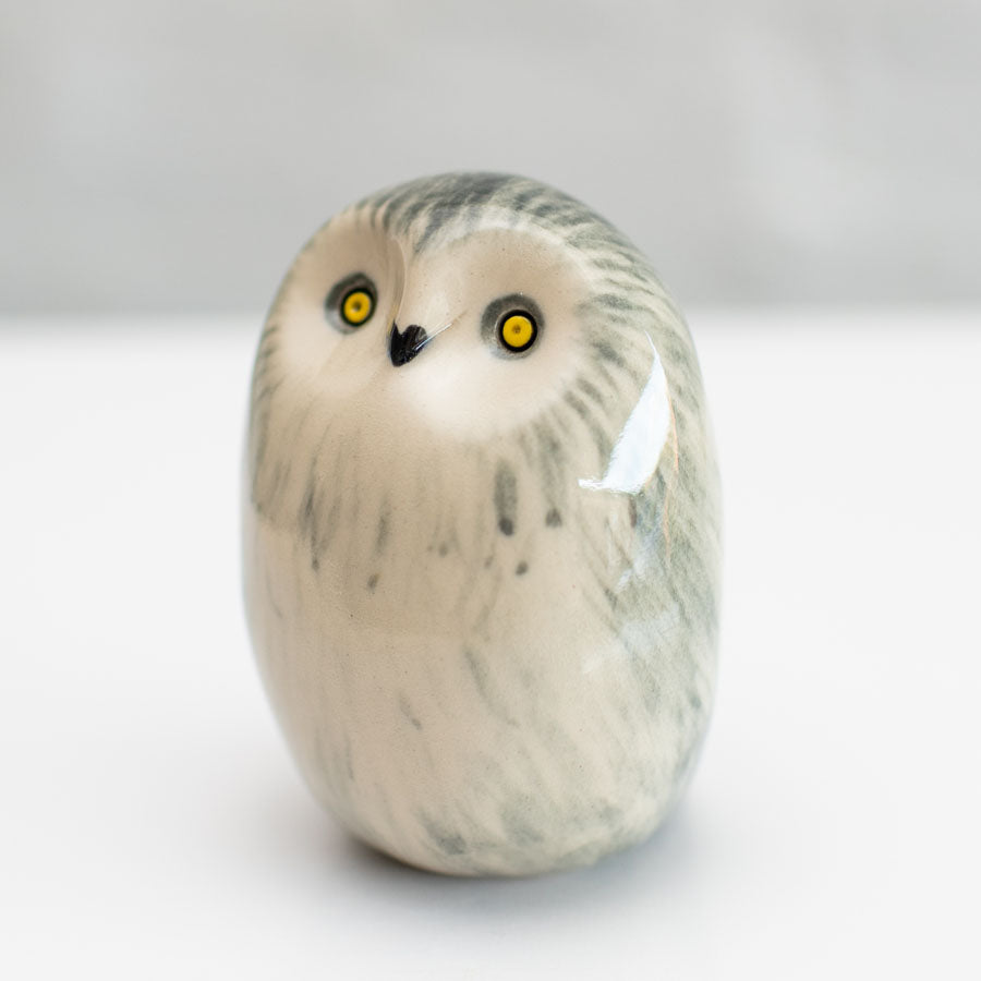 A small sculpted owl figurine with a smooth, rounded body, featuring gray and black feather-like markings on an off-white base, with bright yellow eyes and a small black beak, set against a neutral light gray background.