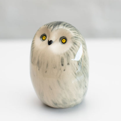 A small sculpted owl figurine with a smooth, rounded body, featuring gray and black feather-like markings on an off-white base, with bright yellow eyes and a small black beak, set against a neutral light gray background.