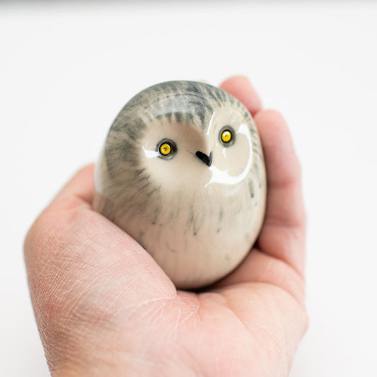 A hand holding a small, rounded owl figurine with gray and white feathers and bright yellow eyes against a plain white background.