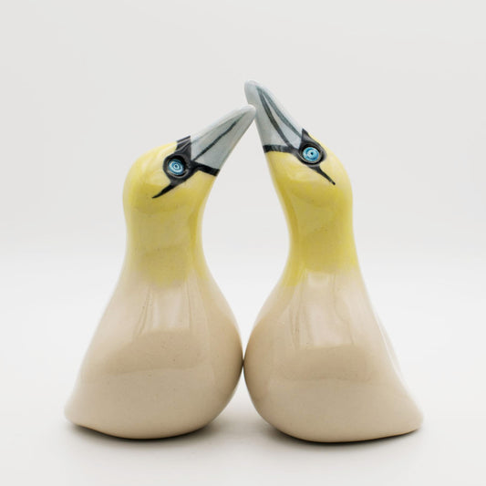 A pair of glossy ceramic Gannet sculptures with pale yellow heads, cream-colored bodies, and gray beaks, facing each other against a plain white background.