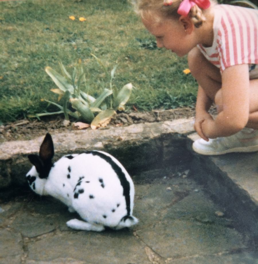 Young girl in a striped shirt crouching to look at a black and white spotted rabbit on a patio