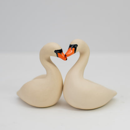 A pair of small white ceramic swan sculptures touching beaks on a plain background