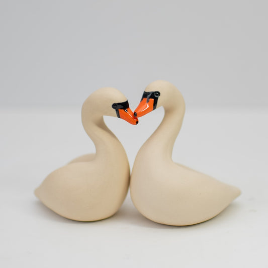 A pair of small white ceramic swan sculptures touching beaks on a plain background