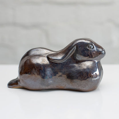 A ceramic hare sculpture with a glossy, metallic brown and silver glaze, resting on a white surface against a light gray brick wall background.