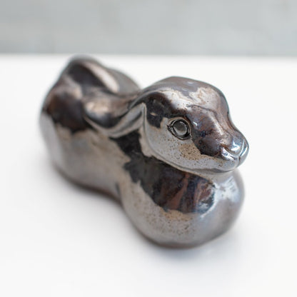 A ceramic hare sculpture with a glossy, metallic brown and silver glaze, resting on a white surface against a light gray brick wall background.