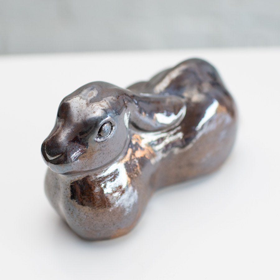 A ceramic hare sculpture with a glossy, metallic brown and silver glaze, resting on a white surface against a light gray brick wall background.