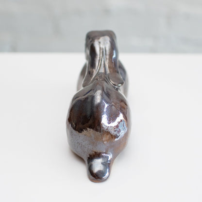 A ceramic hare sculpture with a glossy, metallic brown and silver glaze, resting on a white surface against a light gray brick wall background.