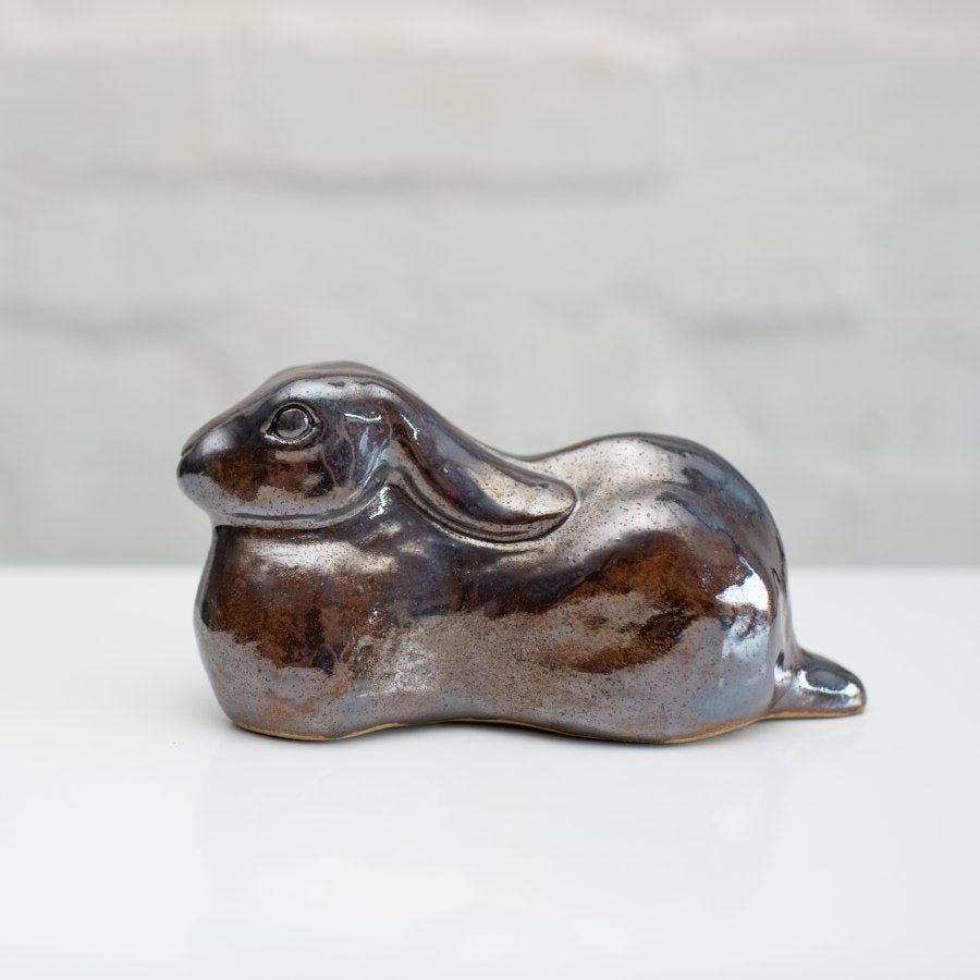 A ceramic hare sculpture with a glossy, metallic brown and silver glaze, resting on a white surface against a light gray brick wall background.