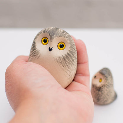 A hand holds a small, rounded owl figurine with yellow eyes and a black beak, with a second owl figurine blurred in the background on a white surface.
