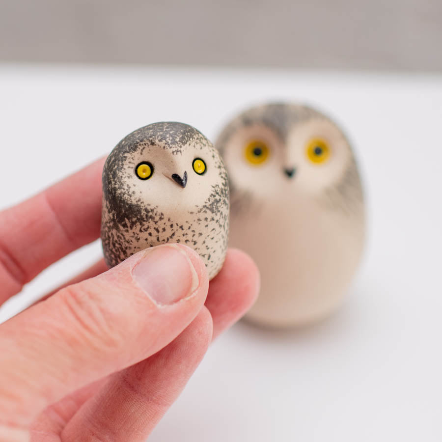 A hand holds a small owl figurine with speckled gray and white feathers and bright yellow eyes, with a second similar owl figurine blurred in the background on a light surface.