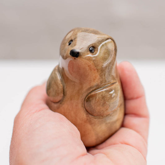 A small glossy ceramic stoat sculpture against a grey backdrop in a hand