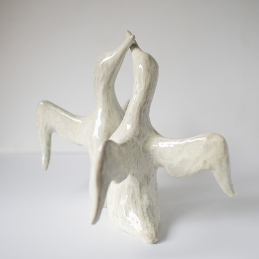 Ceramic sculpture of two abstract birds with long necks and wings, touching beaks, light gray and white glaze