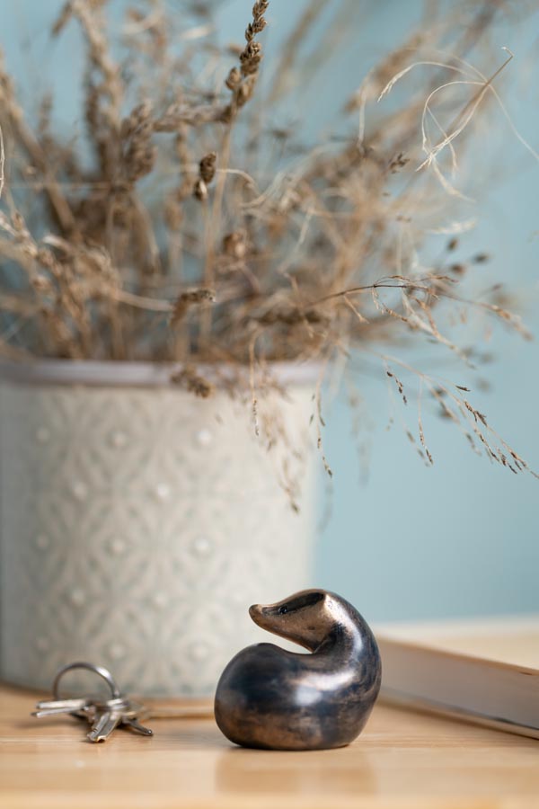 A small bronze badger sculpture on a wooden surface next to keys and a book with a vase of dried grass in the background