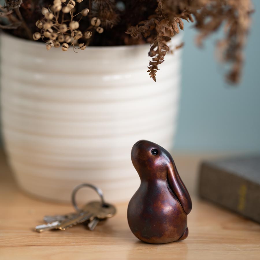Small Brown Foundry Bronze Bunny Rabbit Sculpture on a wooden surface next to keys and a book with dried flowers in a white vase in the background